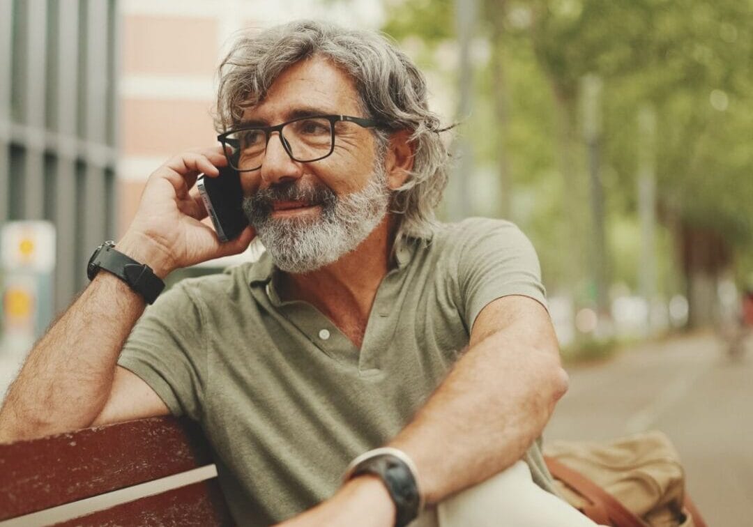 Laughing middle-aged man with gray hair and beard wearing casual clothes sits on bench and uses mobile phone. Mature gentleman in eyeglasses talking on smartphone